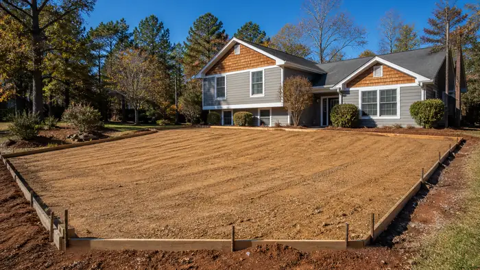 A plate compactor tamps down soil in a small excavated area. at a grey split-level home in Warner Robins, GA, in clear late-morning sun.